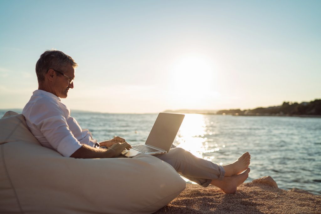 Senior man working on his laptop lying on deck chair on the beach during sunset A Small business owner sitting on a bean bag chair at the beach, using a laptop and feeling satisfied with their business growth.